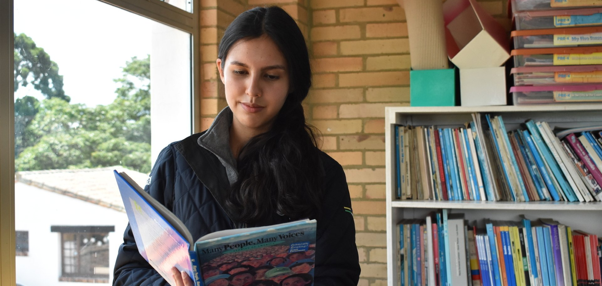 Una persona leyendo un libro colorido junto a una estantería en una habitación luminosa.