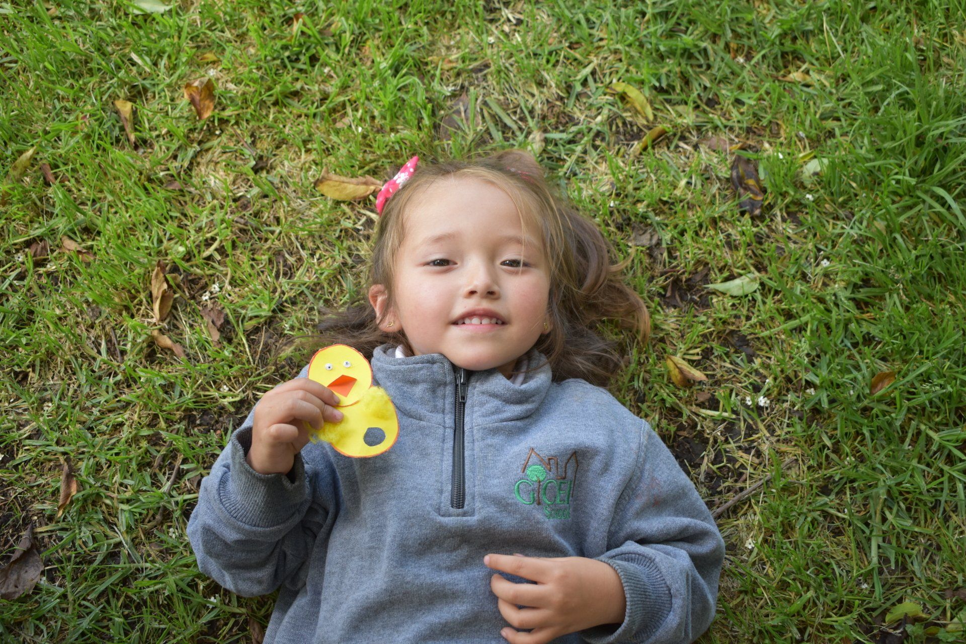 Un niño tumbado en la hierba, sosteniendo un patito de juguete amarillo, con una chaqueta gris y mirando hacia arriba con una leve sonrisa.