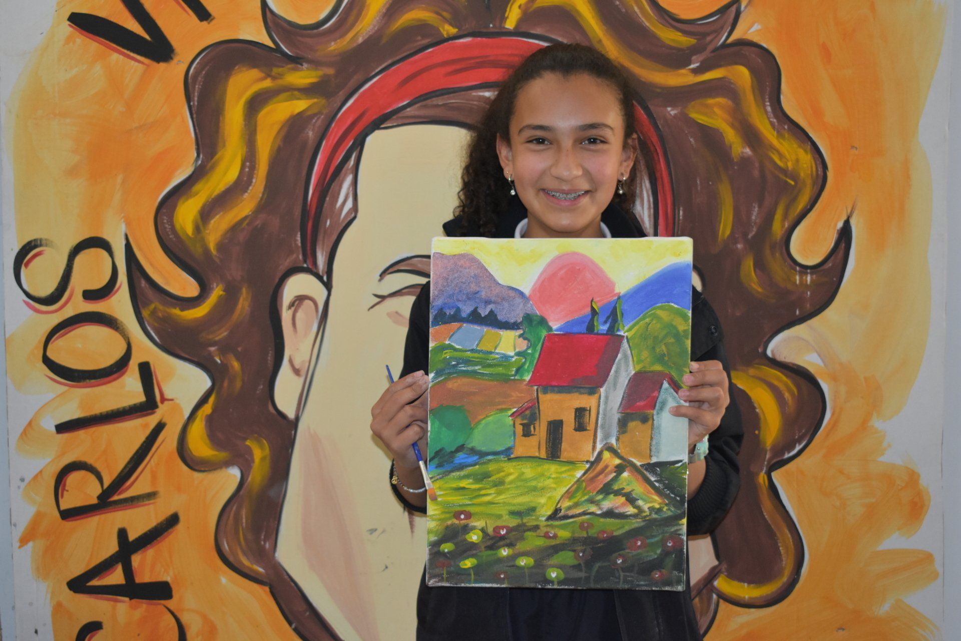 Niña sosteniendo un cuadro colorido frente a un mural naranja con la palabra “Carlos” visible.