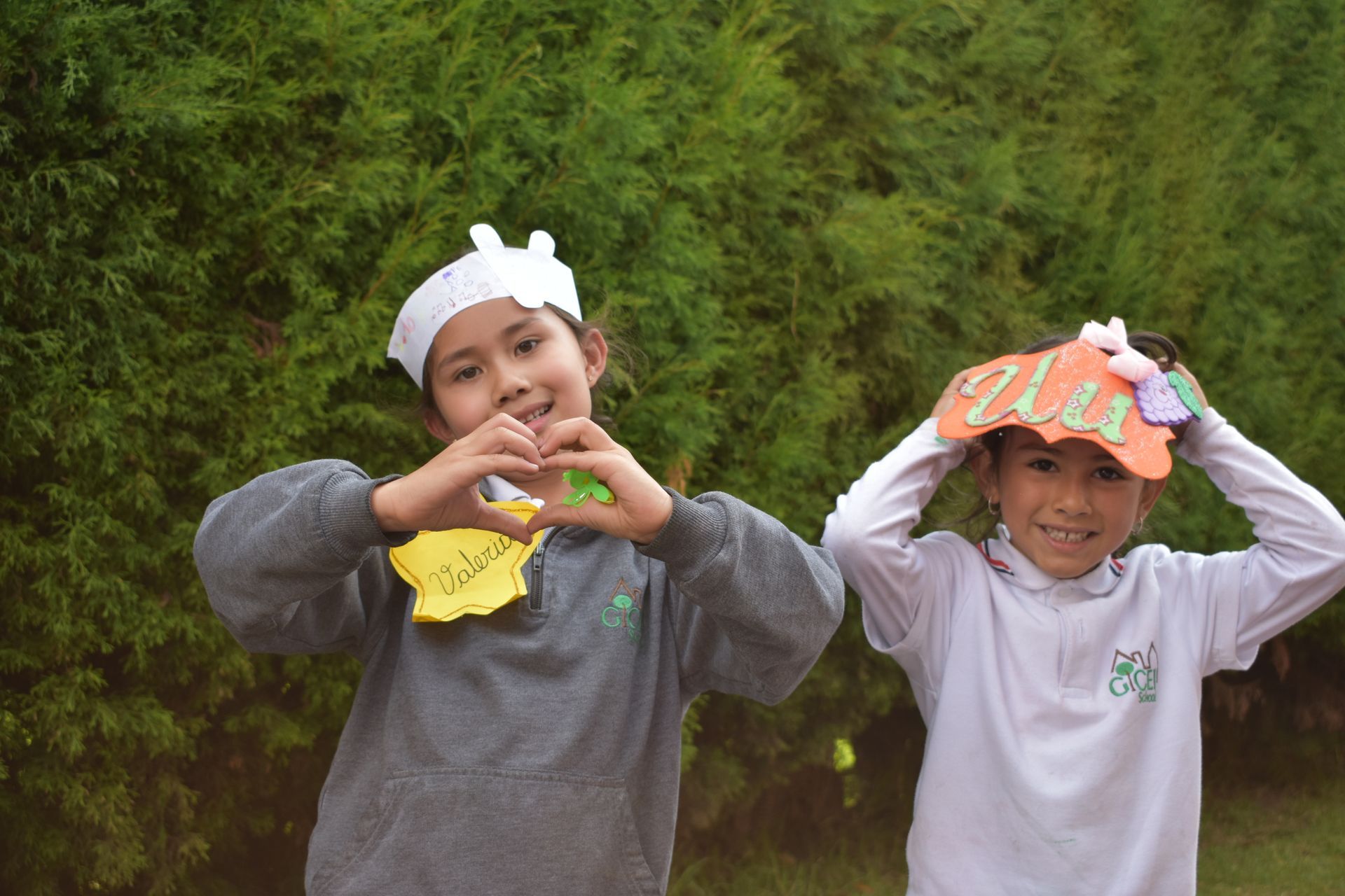 Dos niños con diademas posan delante de un seto verde, formando corazones con las manos.