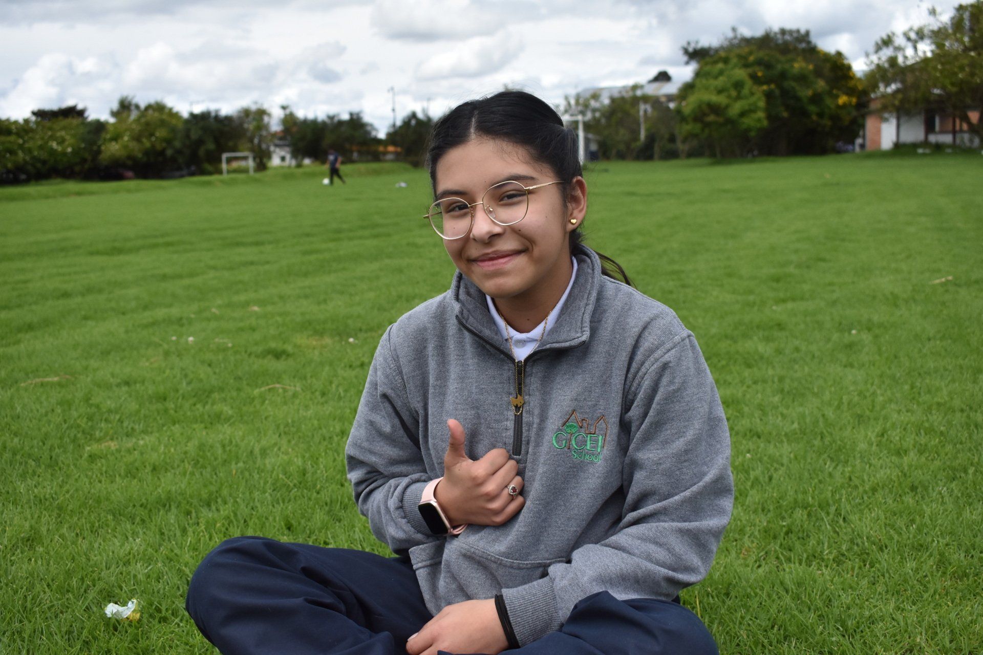 Una persona sentada en el césped de un parque, sonriendo y levantando el pulgar.