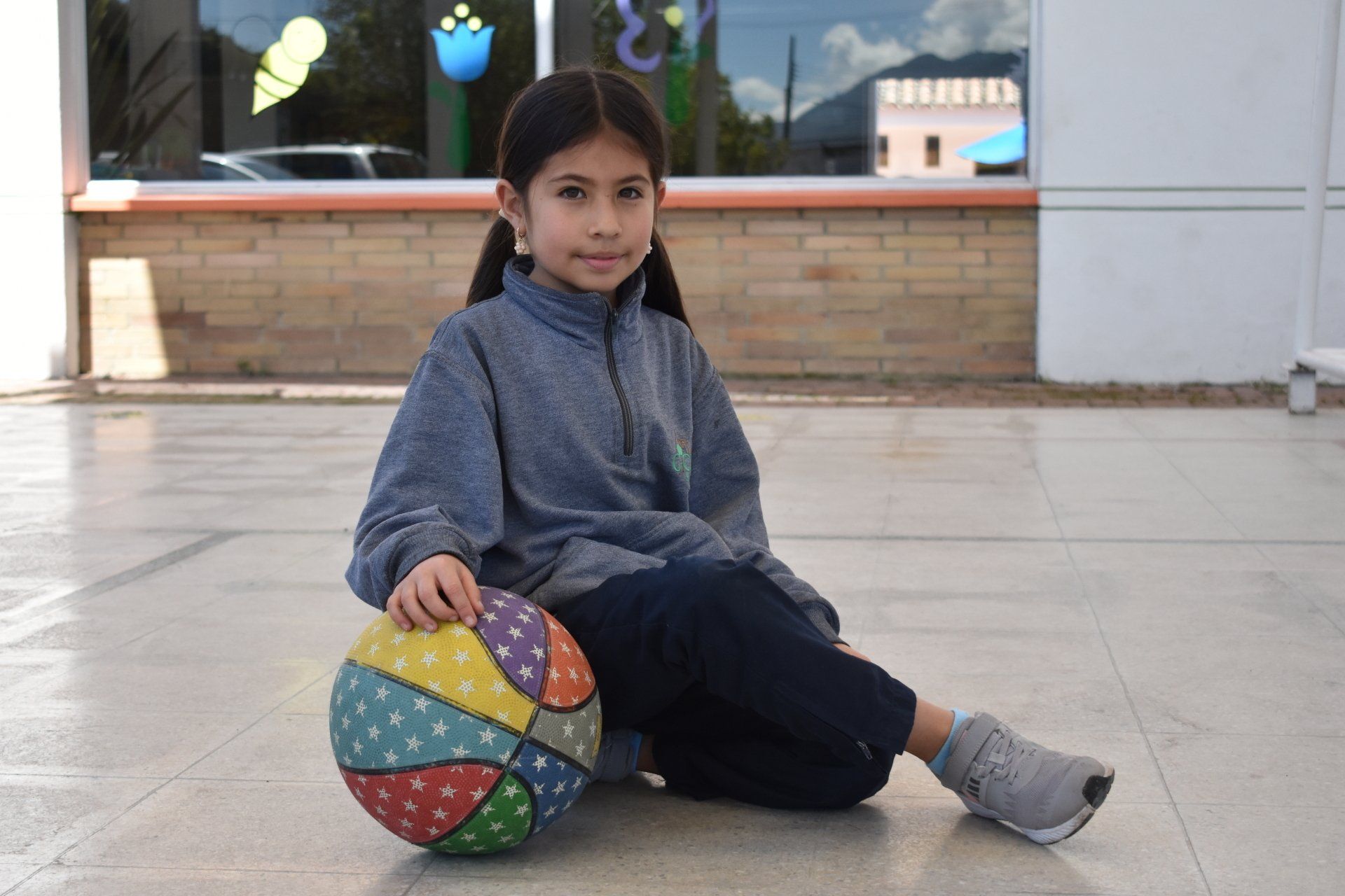 Un niño está sentado en un patio, apoyado en una pelota de colores cerca de la entrada de un edificio.