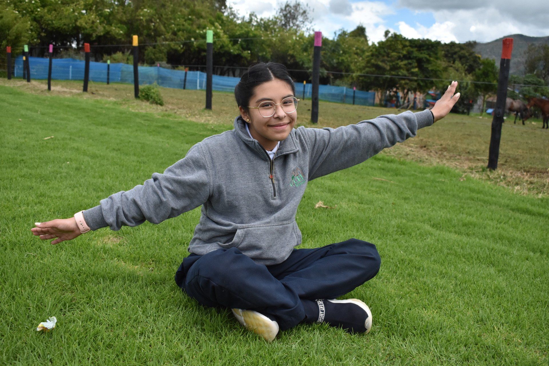 Persona sentada en el césped con los brazos extendidos, sonriendo, vestida con una chaqueta gris y pantalones oscuros, cerca de la valla de un parque.