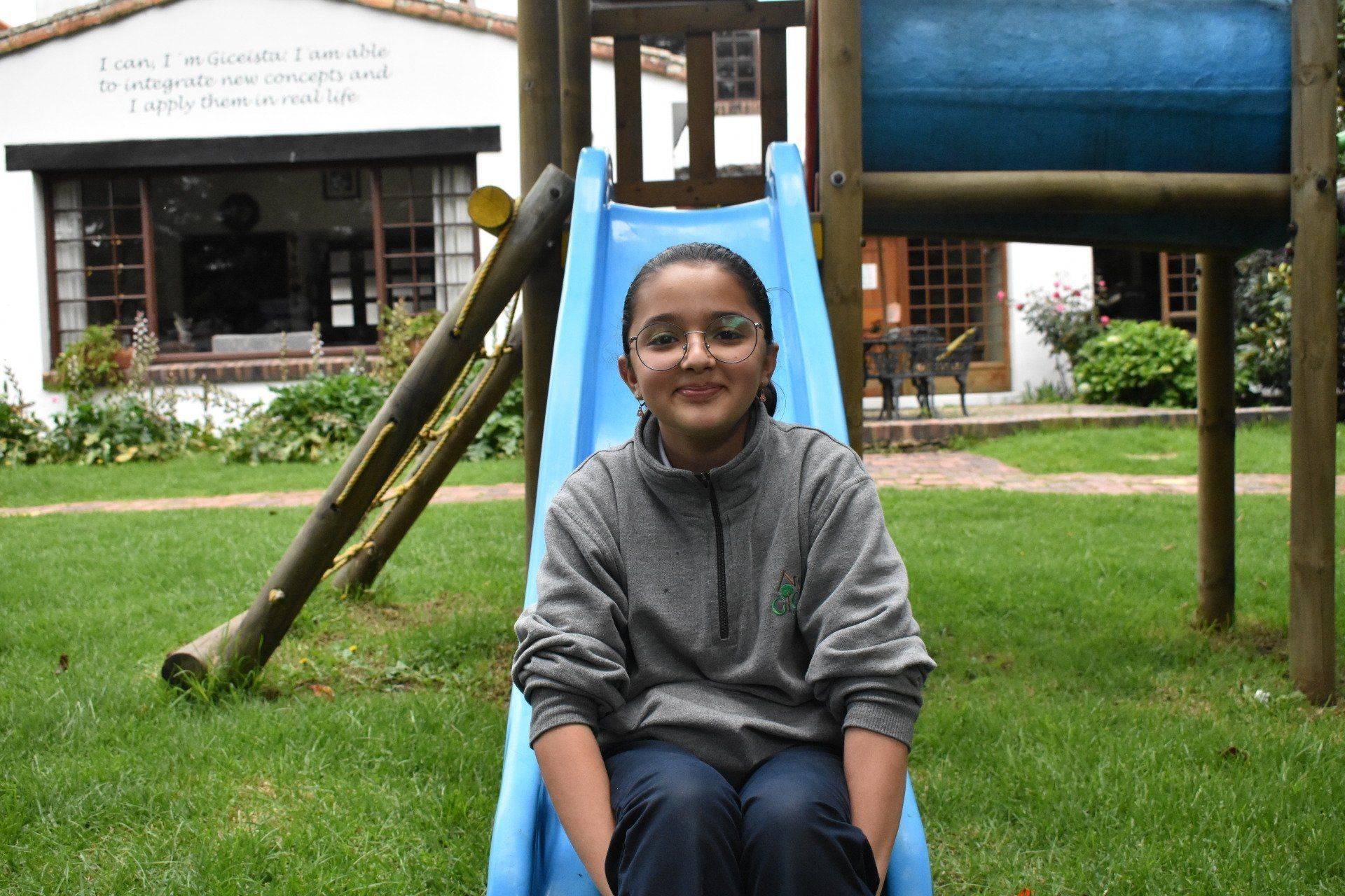 Una persona sentada en un tobogán azul de un parque infantil, sonriendo en un césped cerca de un edificio.