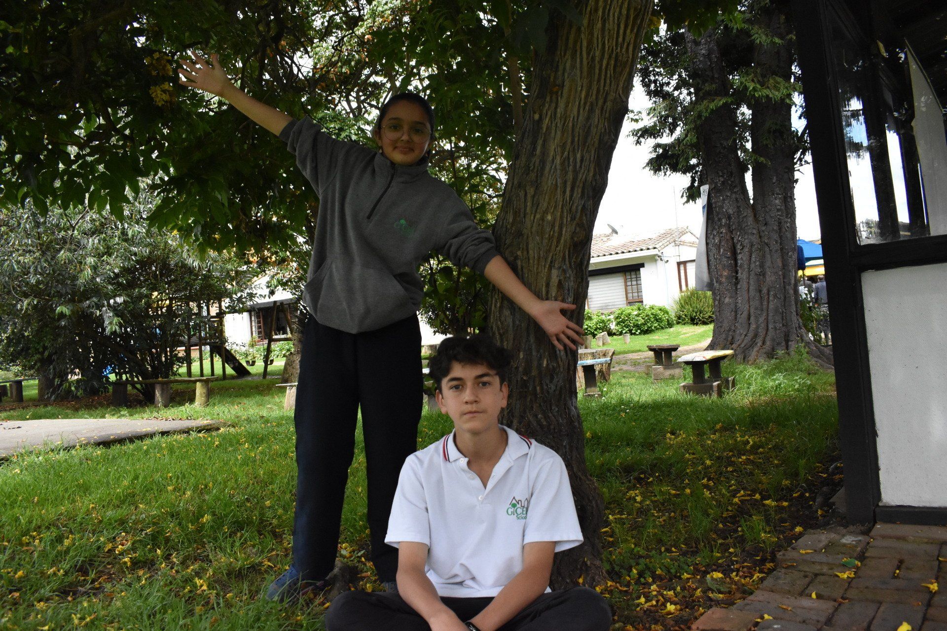 Dos personas posando bajo un gran árbol en un parque; una de ellas está de pie con los brazos extendidos y la otra sentada delante.