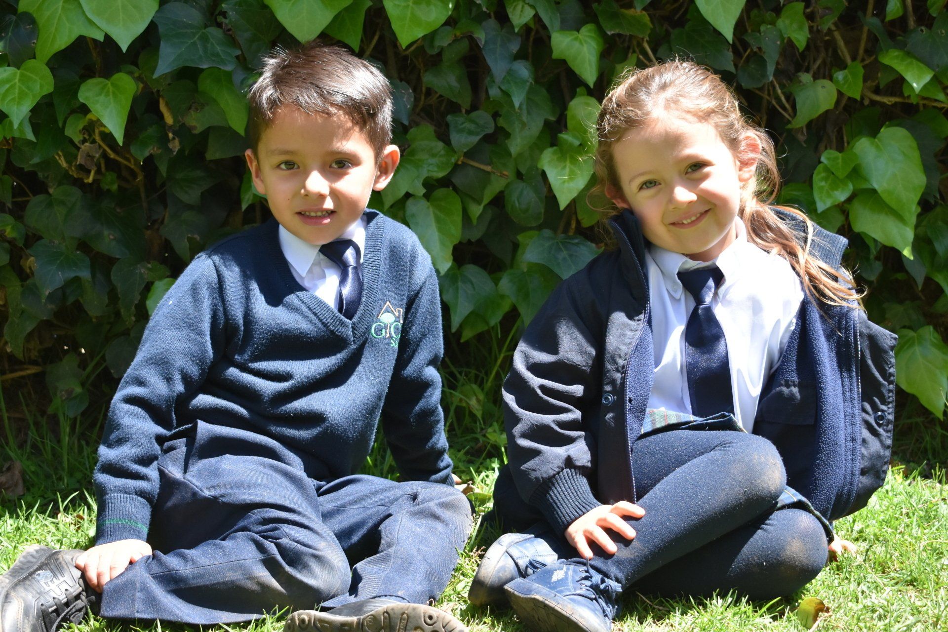 Dos niños sentados en el césped, con uniforme escolar, frente a unos arbustos frondosos.
