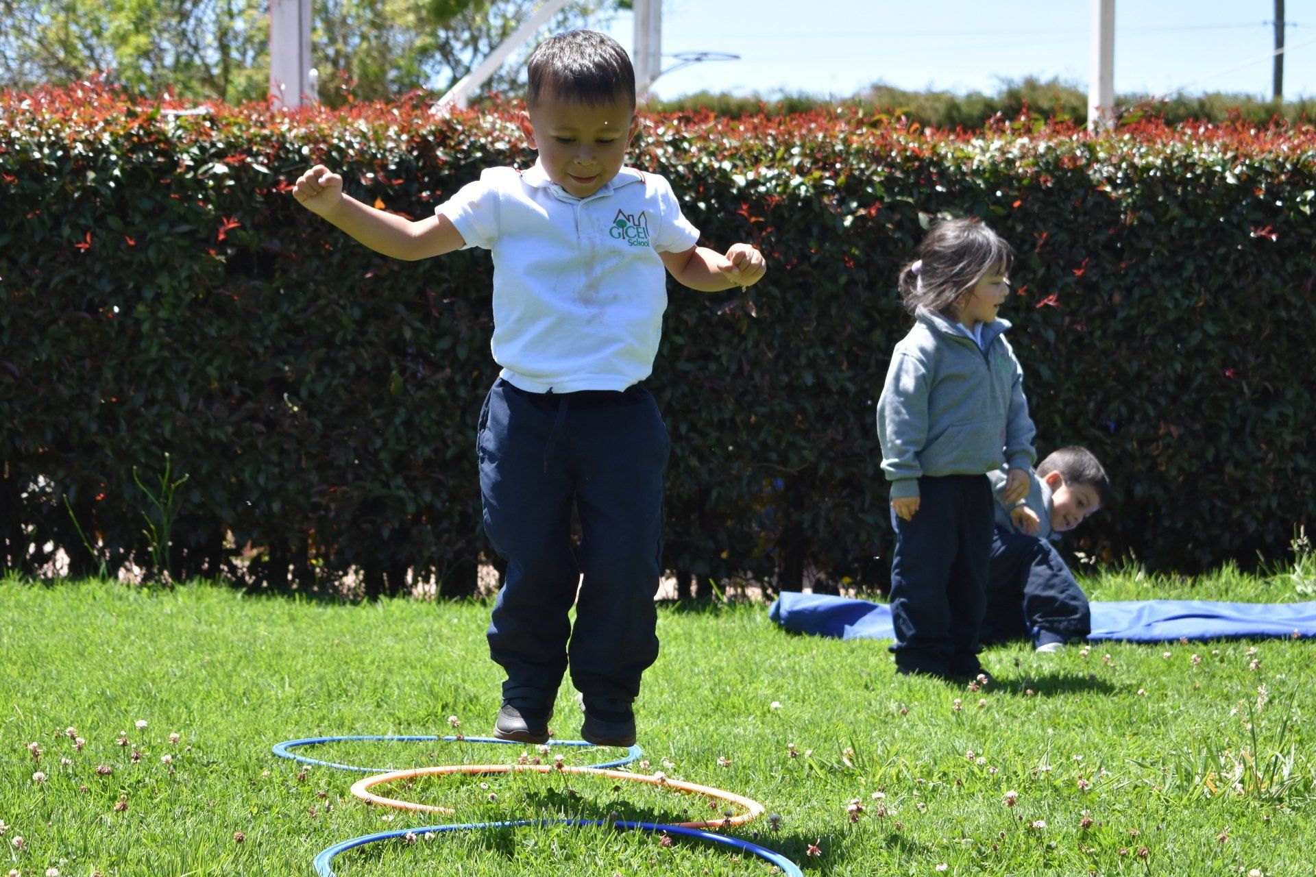 Una niña se equilibra sobre aros en un parque soleado, con dos niños cerca de un seto al fondo.