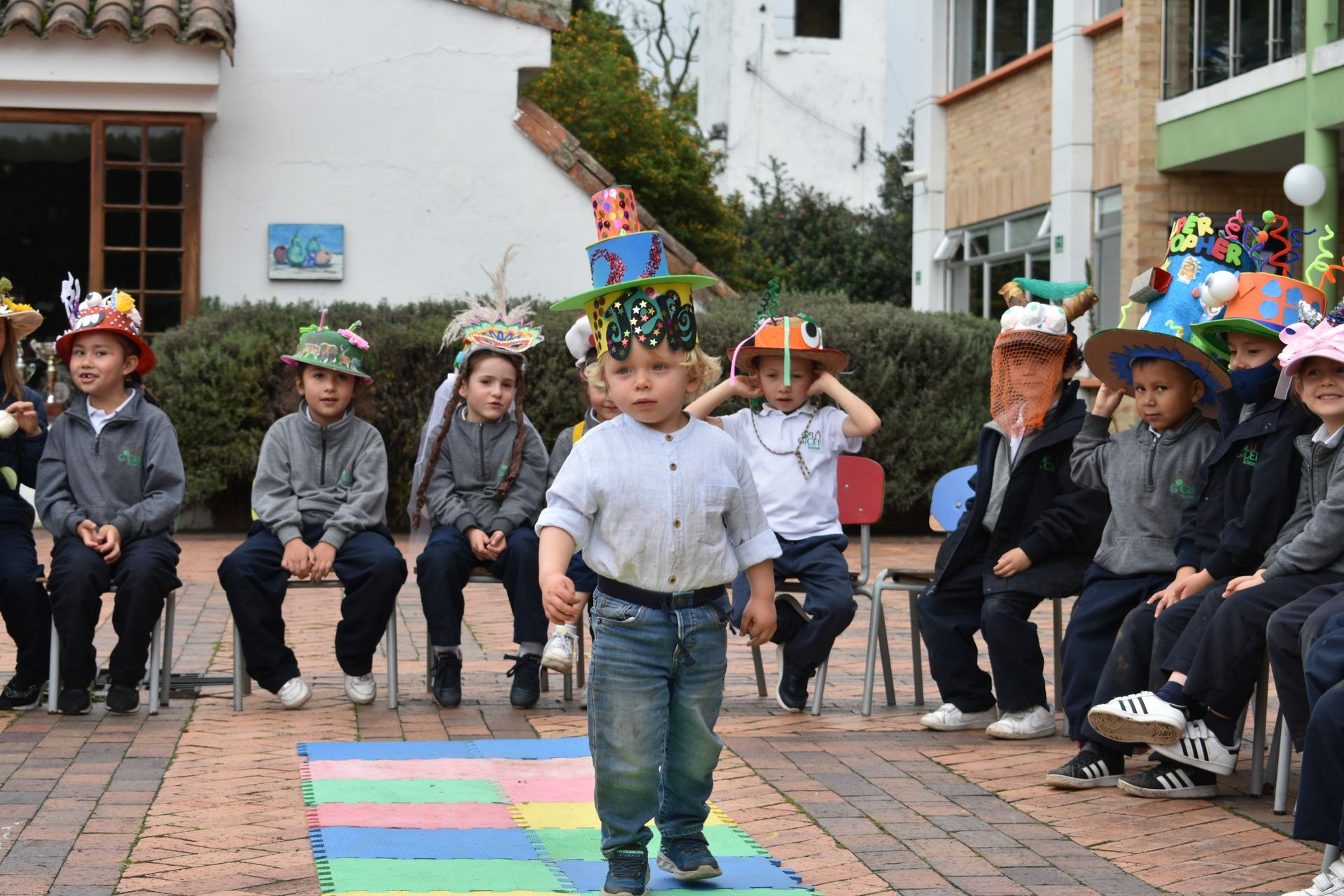 Un niño actúa sobre una alfombra colorida mientras sus compañeros, con sombreros festivos, lo observan sentados en un patio.