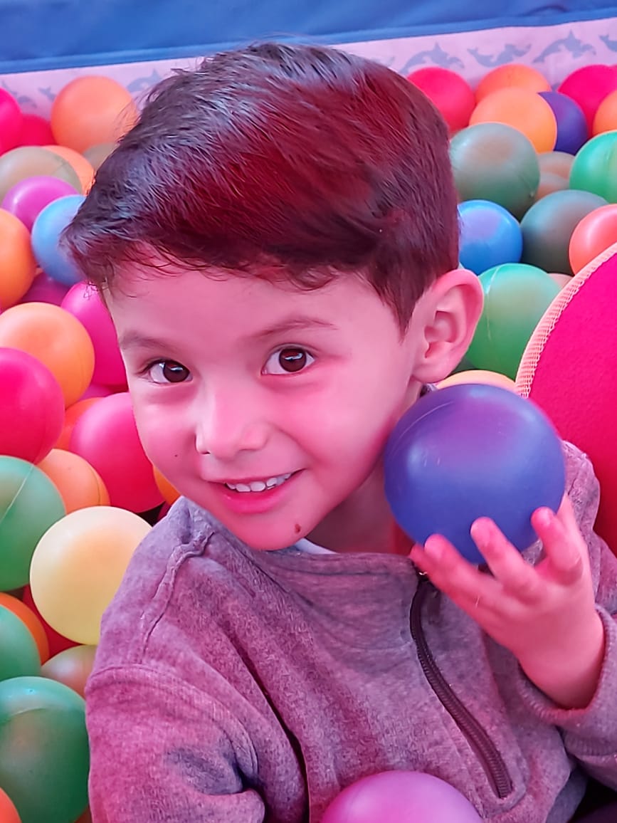 Un niño sonríe en una colorida piscina de bolas, sosteniendo una pelota azul.
