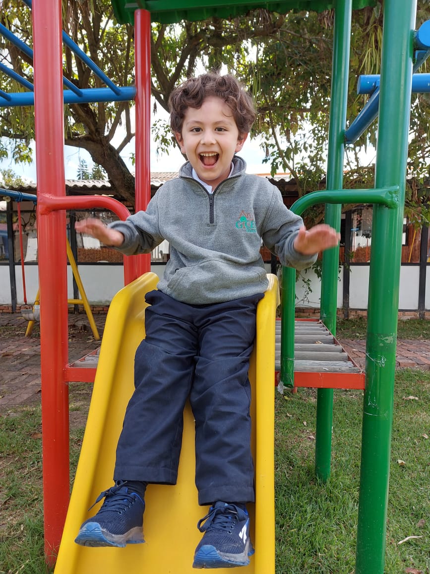 Niño sonriente en un tobogán de un parque infantil, con suéter gris y pantalones azules.