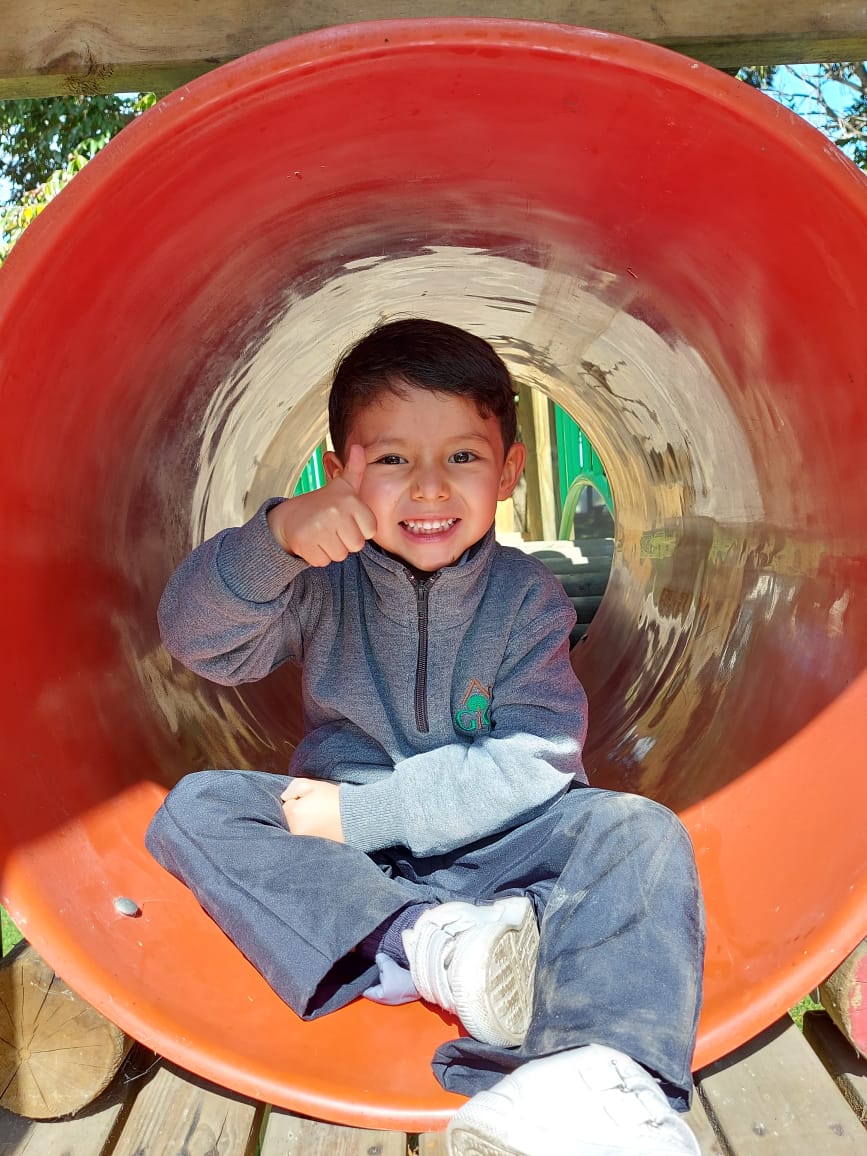 Un niño sonríe sentado dentro de un tobogán tubular rojo en un parque infantil, con una mano en la oreja.