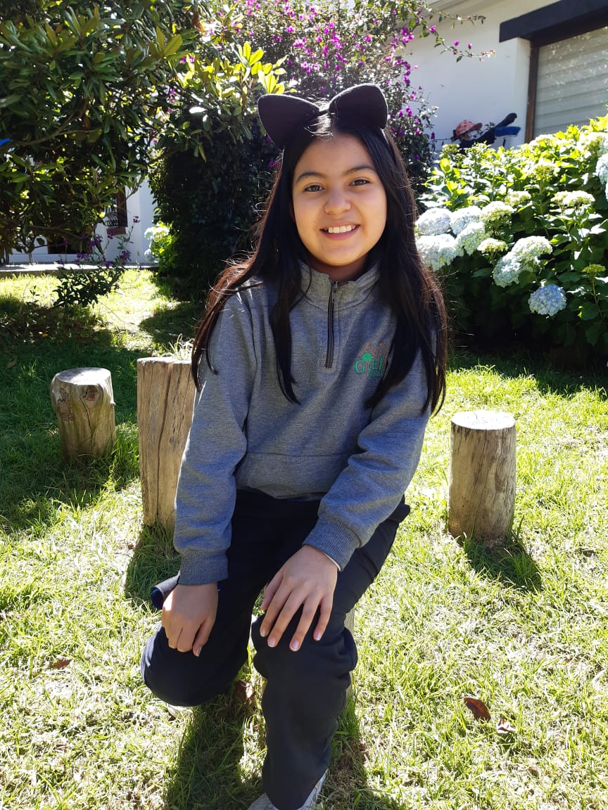 Una persona arrodillada sobre la hierba en un jardín soleado, sonriendo, con flores blancas y árboles detrás.