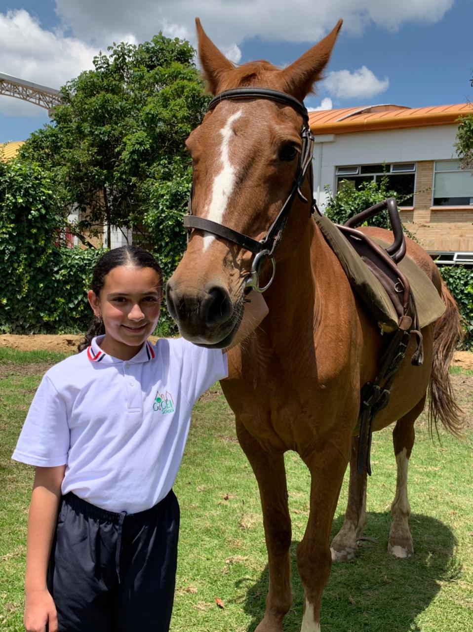 Una niña de pie junto a un caballo marrón sobre la hierba frente a una casa, ambos mirando a la cámara.