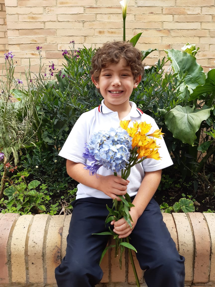 Un niño sonriente sostiene flores azules y amarillas mientras está sentado en un banco de piedra en un jardín.
