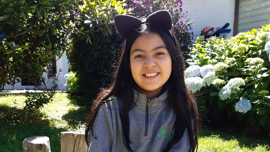 Una persona sonriente con una diadema negra con orejas de gato está de pie en un jardín soleado con flores y vegetación.
