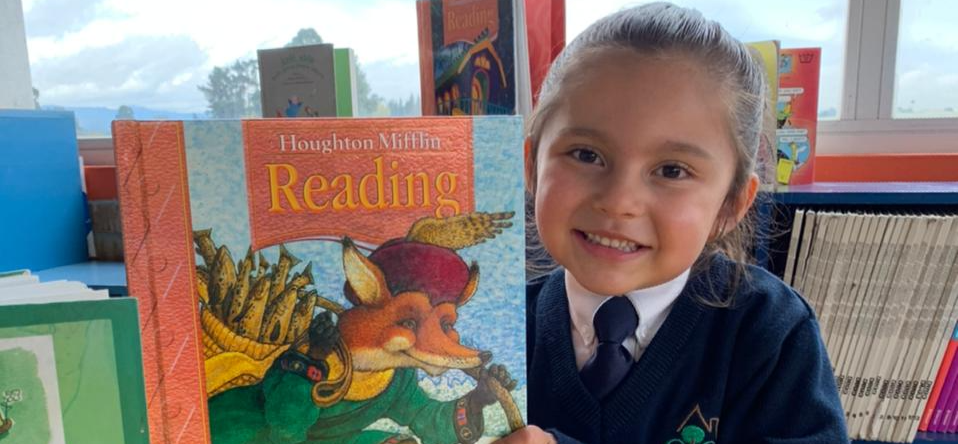 Un niño sonriente sostiene un libro de lectura colorido en el interior de una casa, cerca de una ventana.