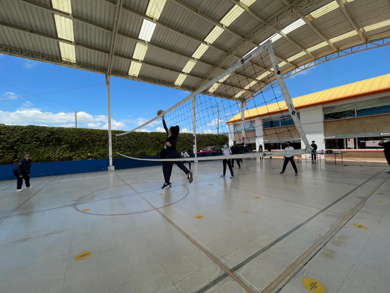 Gente jugando al voleibol en una cancha techada al aire libre cerca de un edificio escolar.