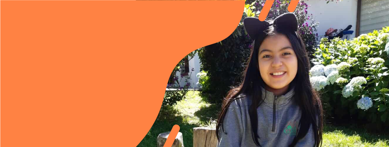 Una persona sonriente con una chaqueta gris está de pie al aire libre junto a unos arbustos en flor y una pared naranja.