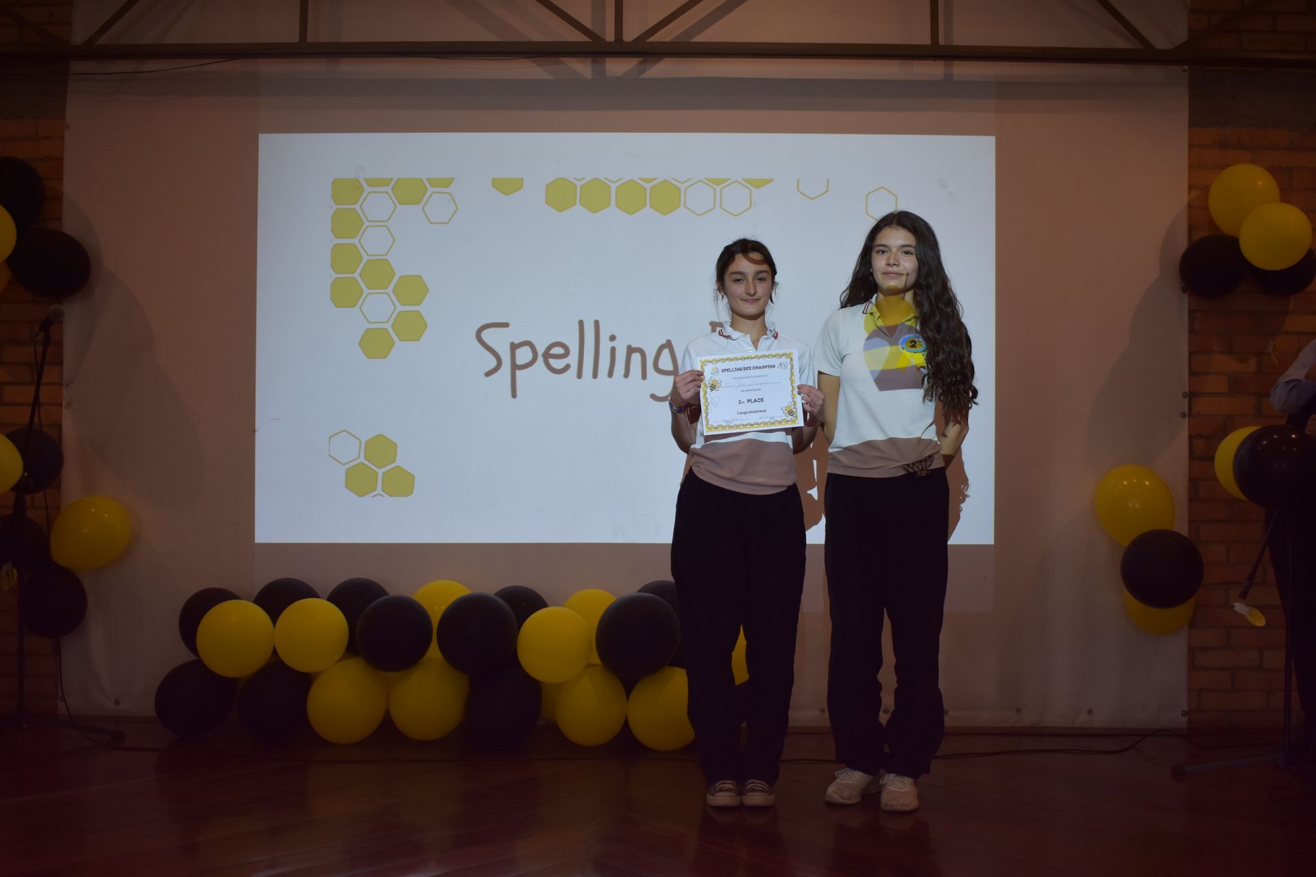 Dos estudiantes sosteniendo certificados en un escenario con un telón de fondo con temática de ortografía y globos amarillos y negros.