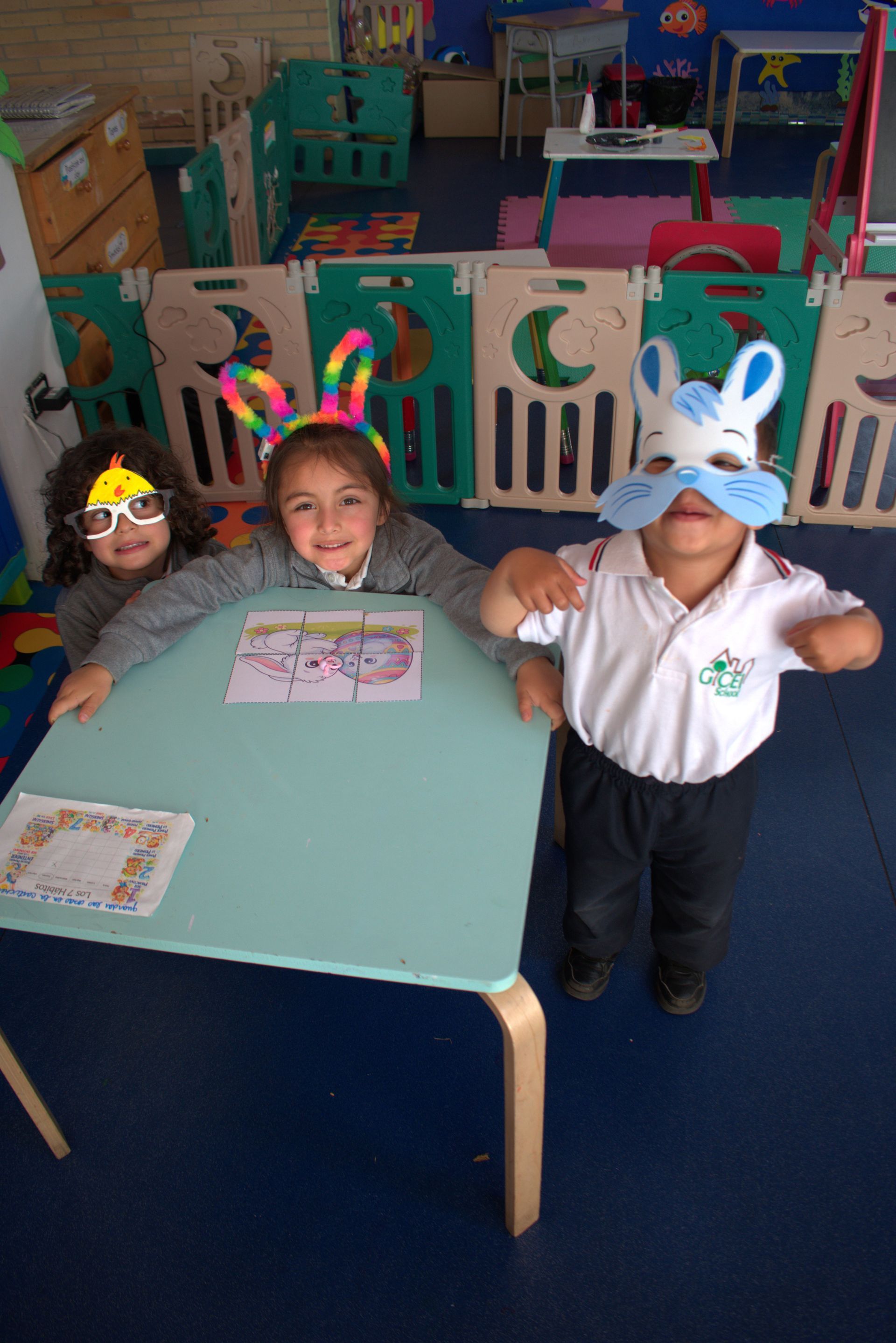 Tres niños con máscaras de conejo están sentados en una mesa de aula con coloridos recortes y hojas de trabajo.
