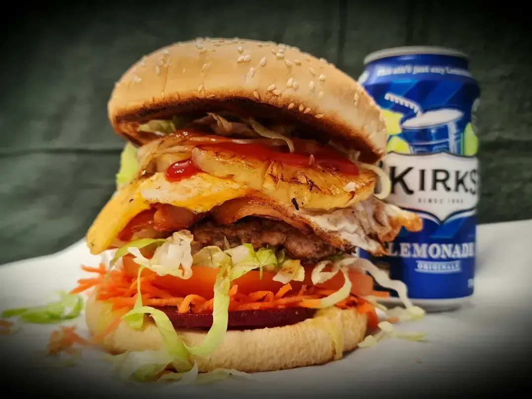 A Tall Burger with Lettuce, Tomato, Pineapple, and Cheese Next to A Can — Sav's Takeaway In Mount Isa, QLD