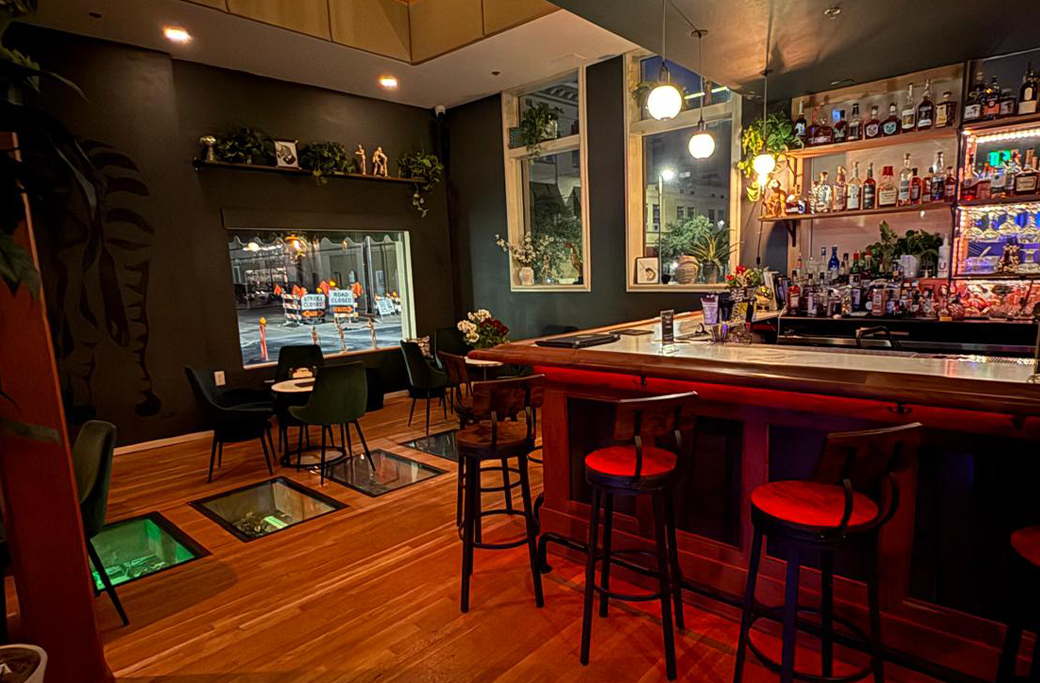 Bar interior with red bar, stools, wooden floor, and a window overlooking a city street at night.