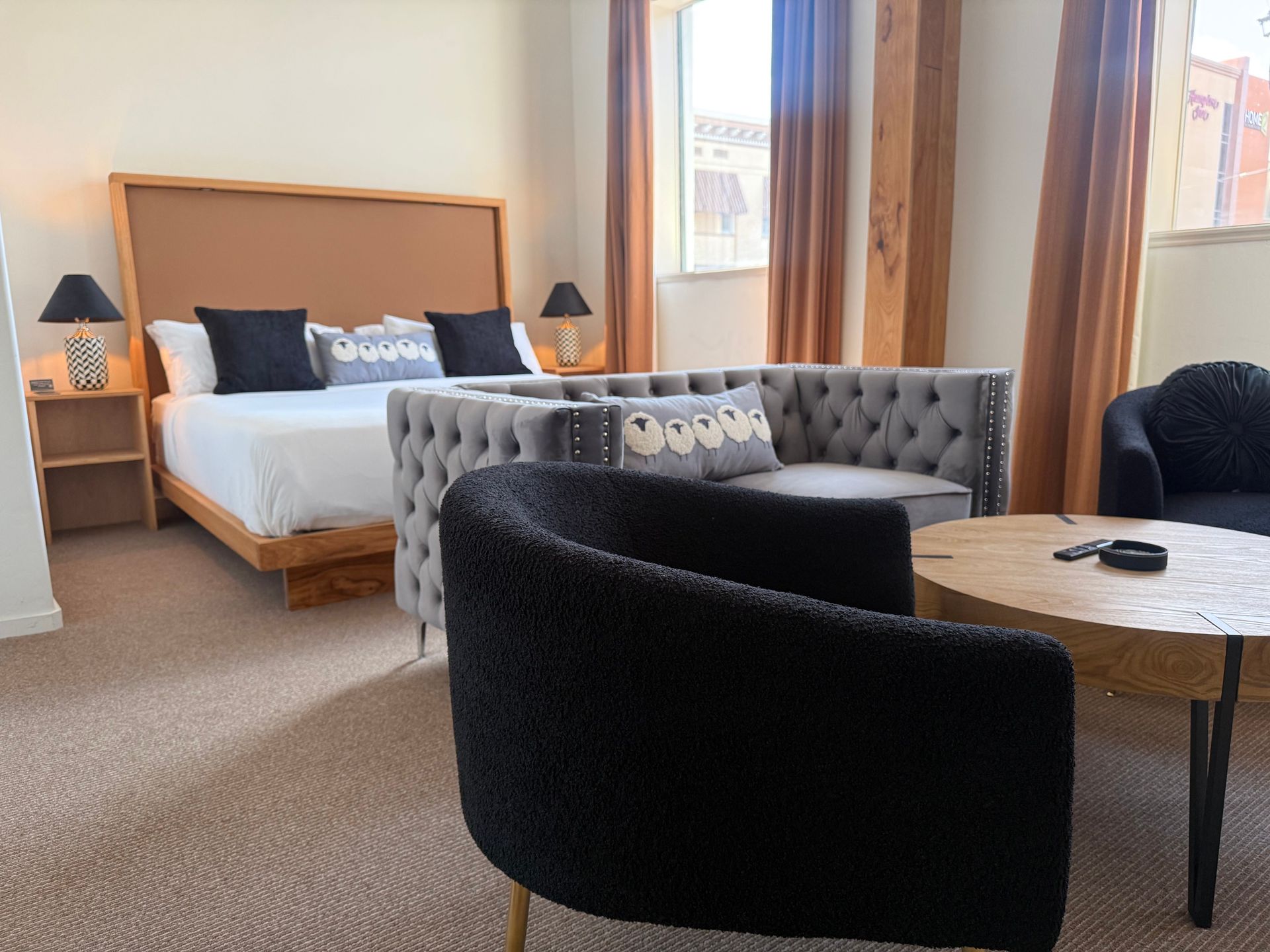 Hotel room with bed, sofa, TV, and large windows with curtains. Neutral tones, well-lit.