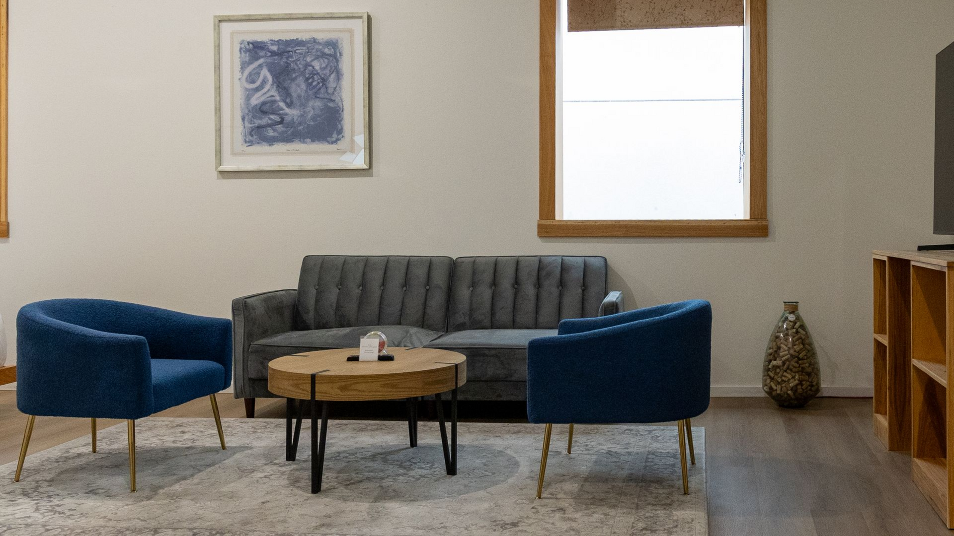 Bedroom with a bed, two blue armchairs, a round coffee table, a clock, and a window.