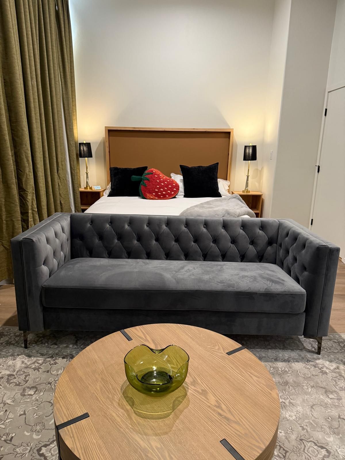 Gray tufted sofa in front of a bed with pillows. Round wooden coffee table in foreground.