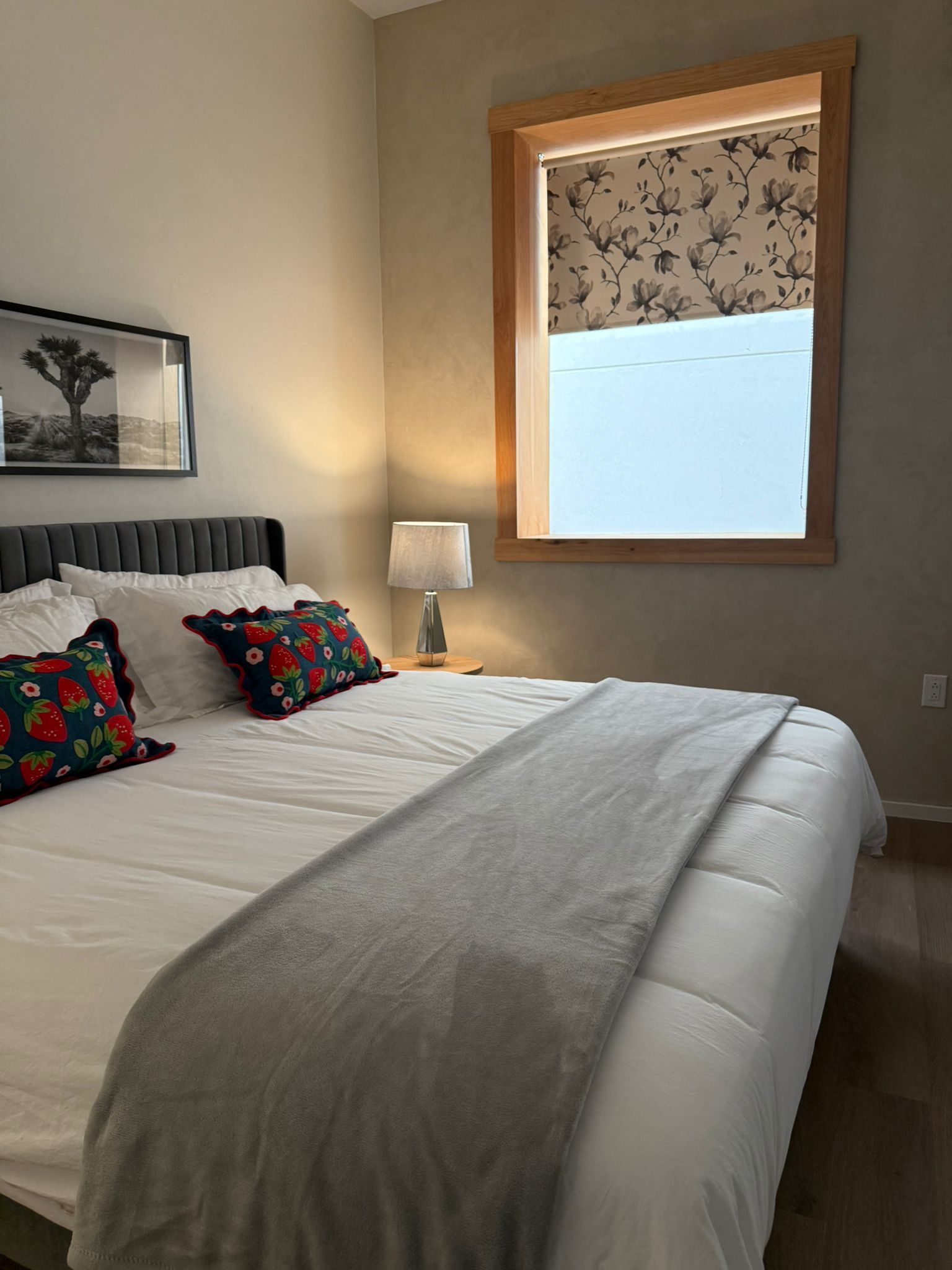 Bedroom with bed, pillows, window, lamp, artwork, and gray blanket.