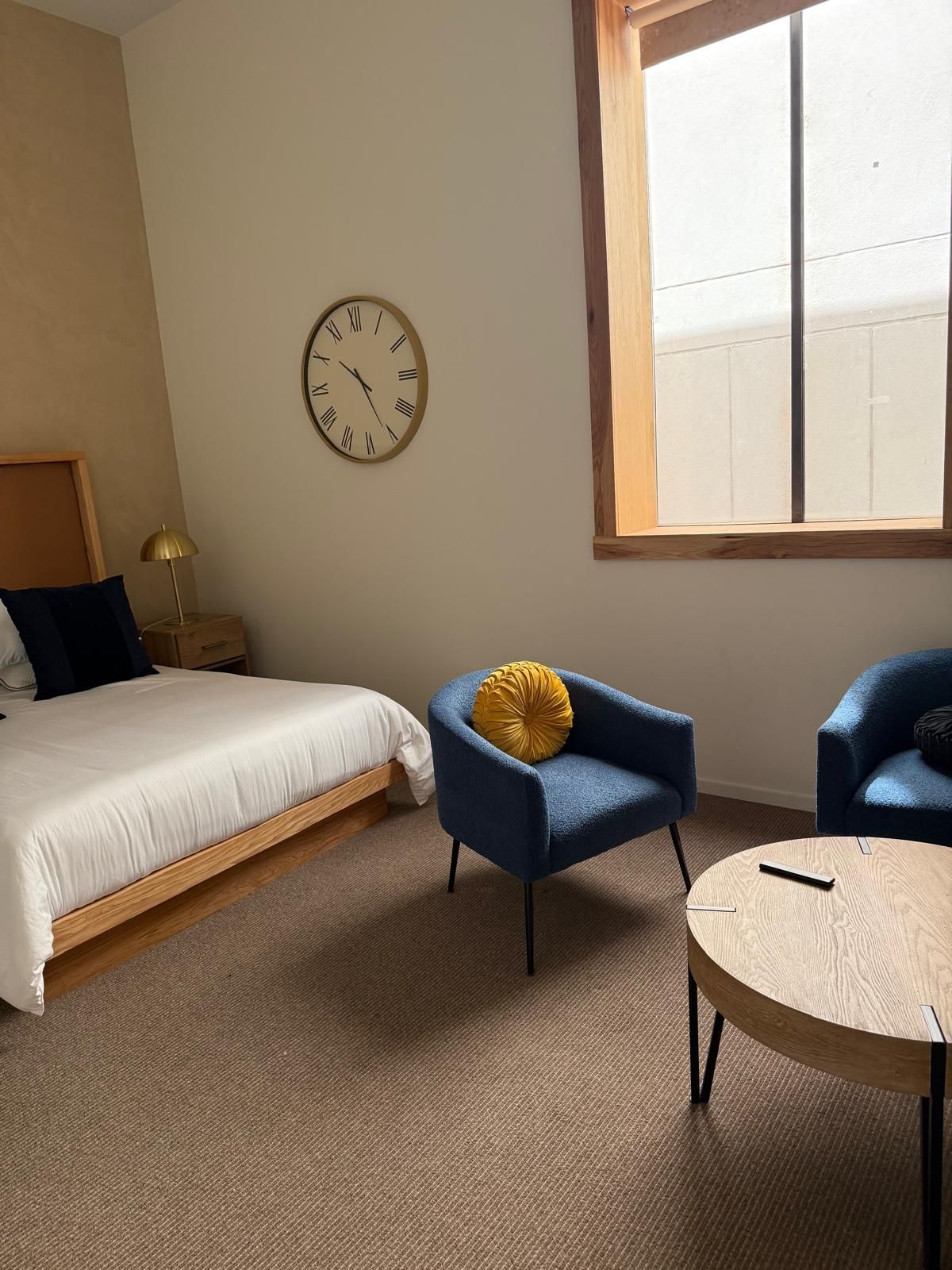 Bedroom with a bed, two blue armchairs, a round coffee table, a clock, and a window.