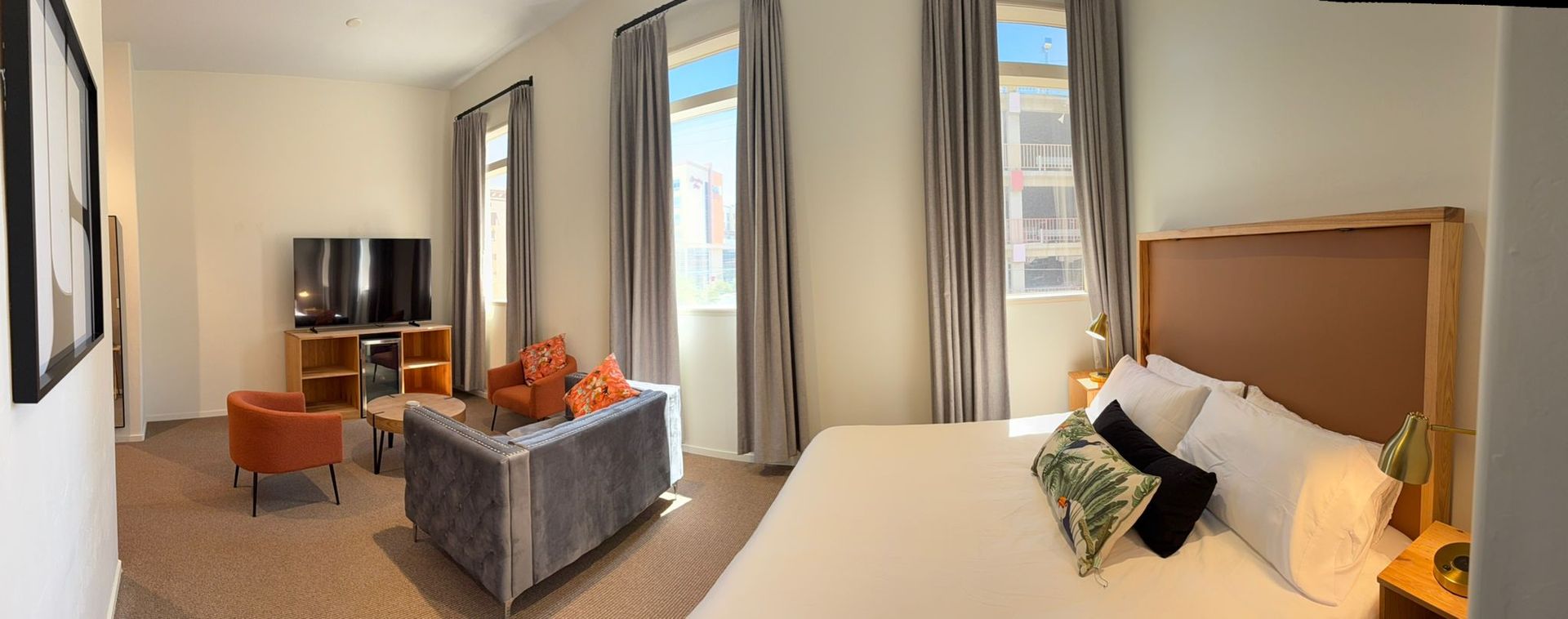 Hotel room with bed, sofa, TV, and large windows with curtains. Neutral tones, well-lit.