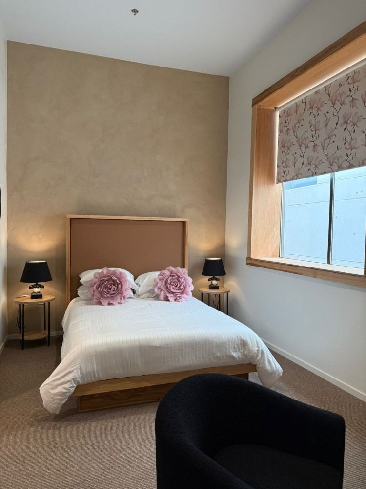Bedroom with a bed, two nightstands, a window, and a black chair. Pale pink and tan color scheme.