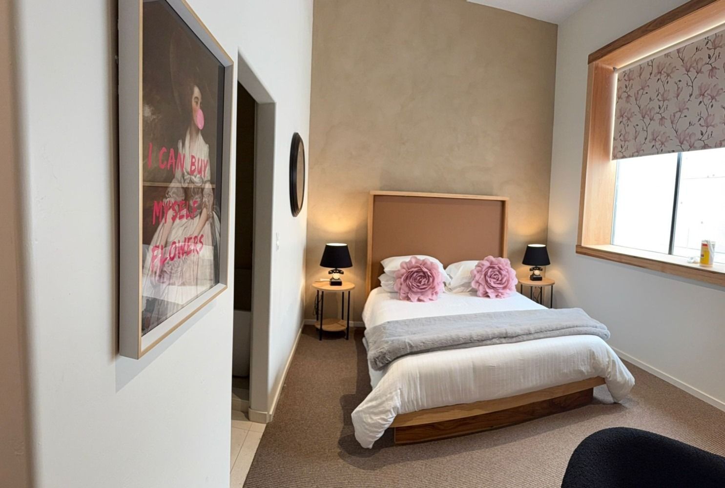 Cozy bedroom with bed, two nightstands with lamps, artwork, and window with patterned blinds.