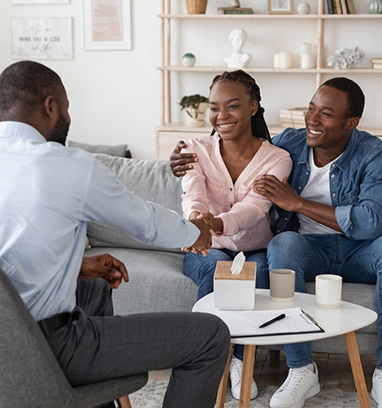 Three people chatting and smiling on a couch in a bright living room, with coffee cups on a table.