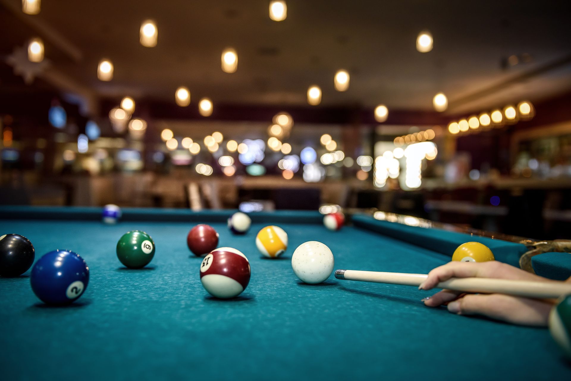 Pool table with multiple colored balls, player's hand with cue stick ready to strike. Dim lighting.