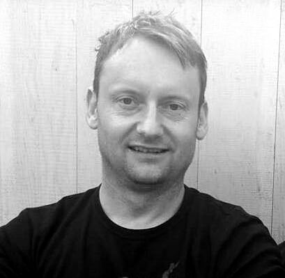 Man smiling, wearing a black t-shirt, in front of a light-colored wood panel background.