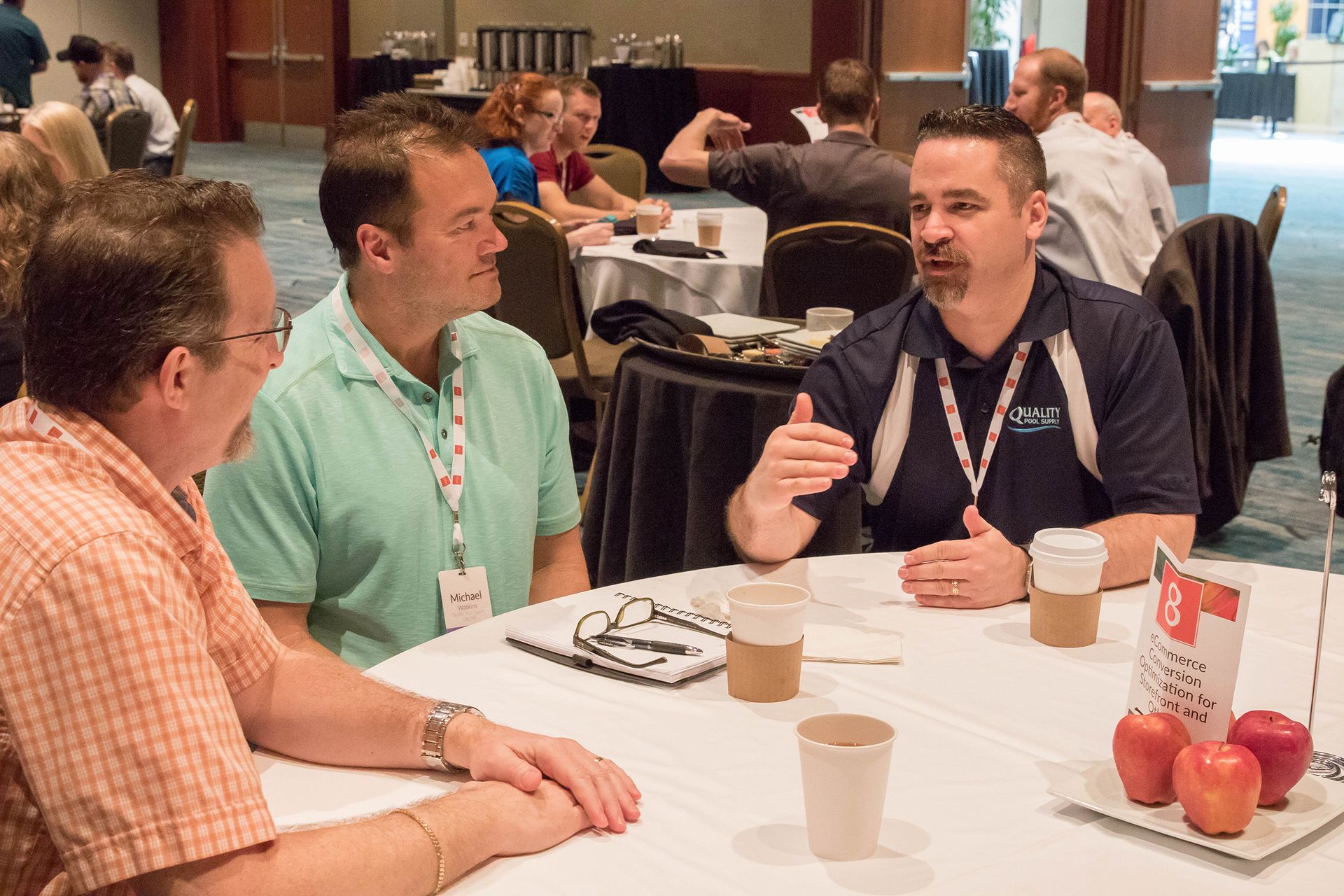 Three men seated at a table in a conference setting, talking. One gestures with his hands, others listen.