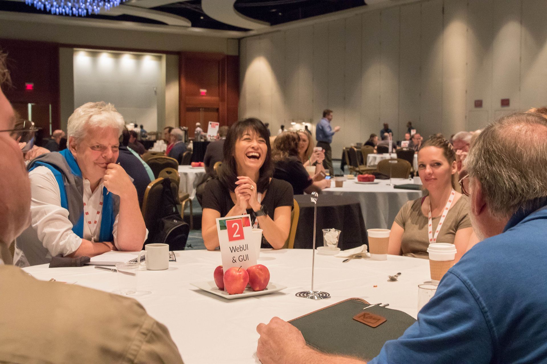 People at round tables in a conference hall, smiling and talking. A woman in the center laughs, clapping hands.