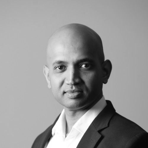 Bald man in a black suit jacket and open white shirt, looking at the camera. Black and white photo.