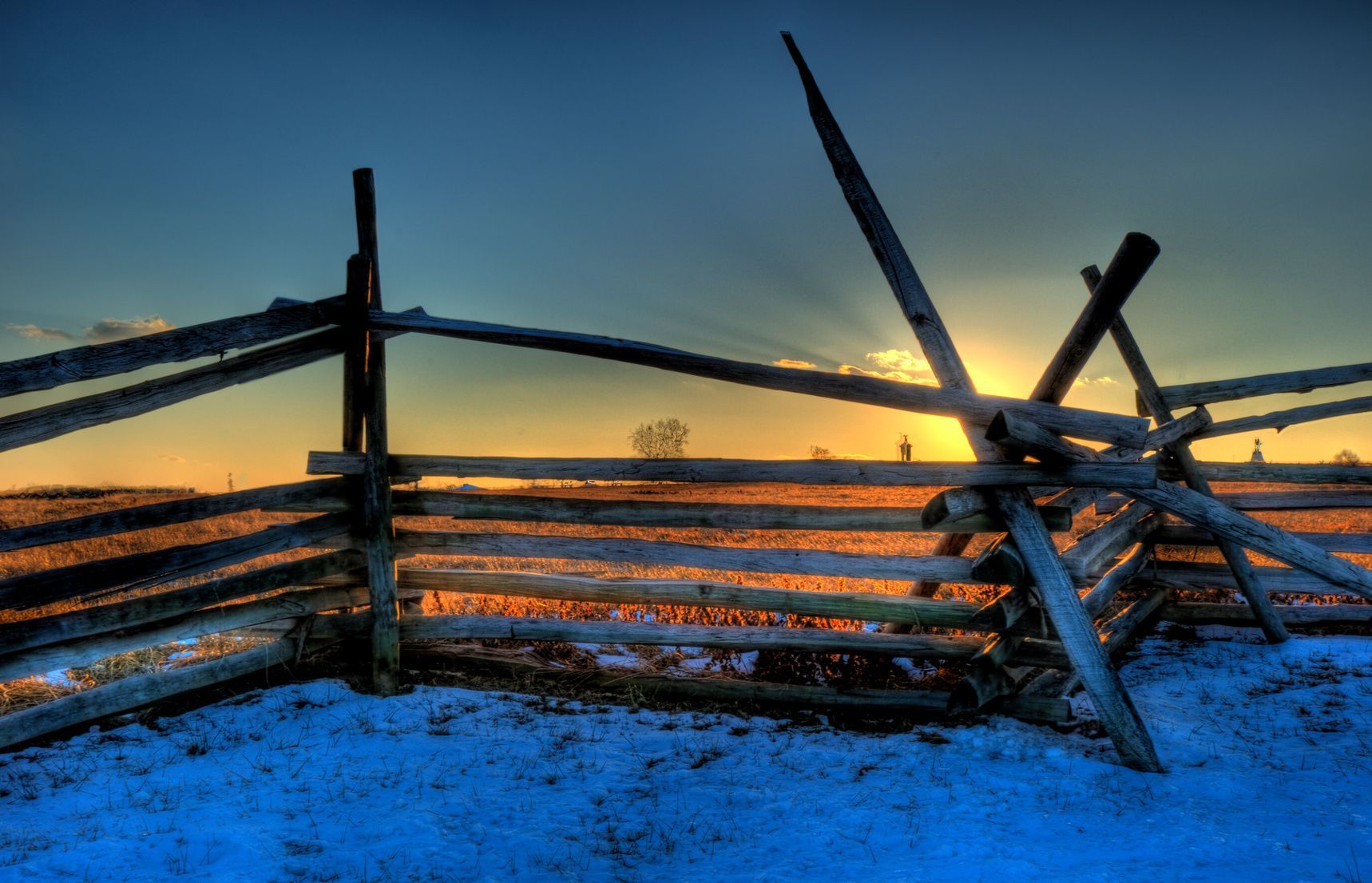 Wooden fence against a sunset, snow-covered ground, orange and blue hues.