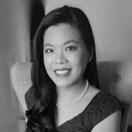Woman with dark hair smiles, wearing a pearl necklace and dark dress, sitting in a chair.
