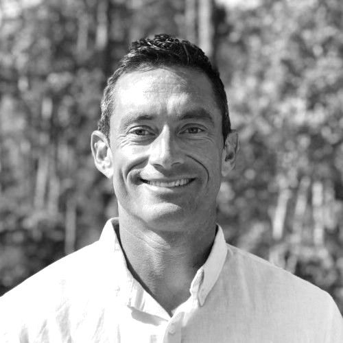 Man smiling outdoors, wearing a light-colored button-down shirt. Trees in the background.