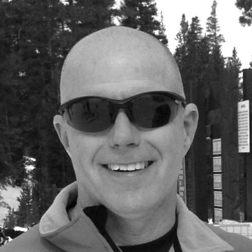 Man with shaved head wearing sunglasses, smiling outdoors.