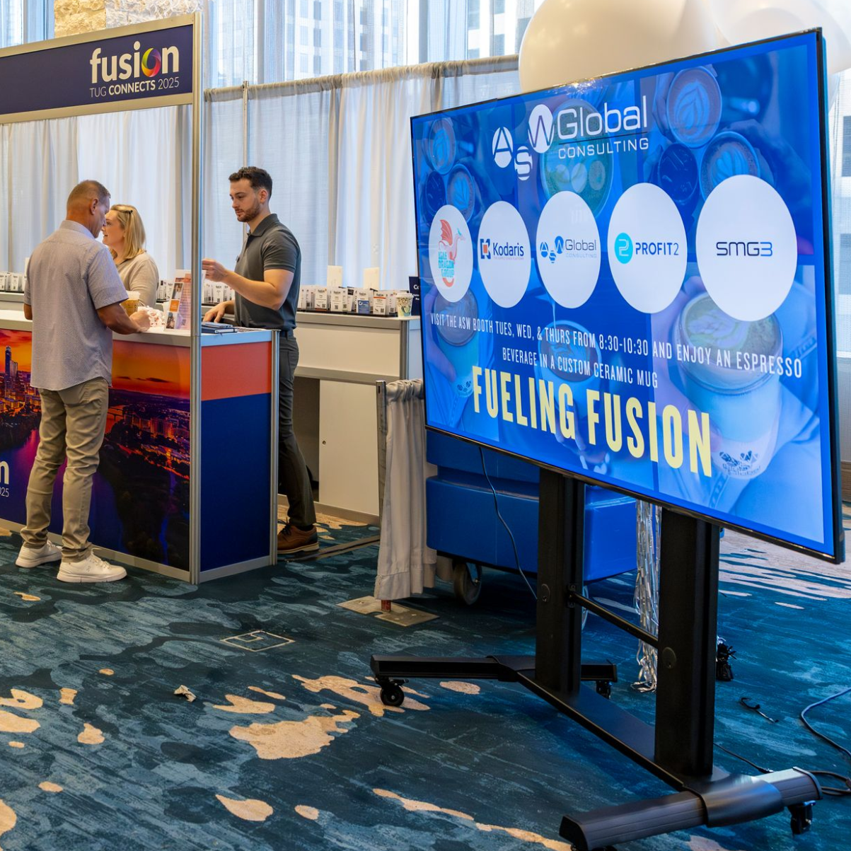 A trade show booth with a large screen displaying logos. People interact at a counter.