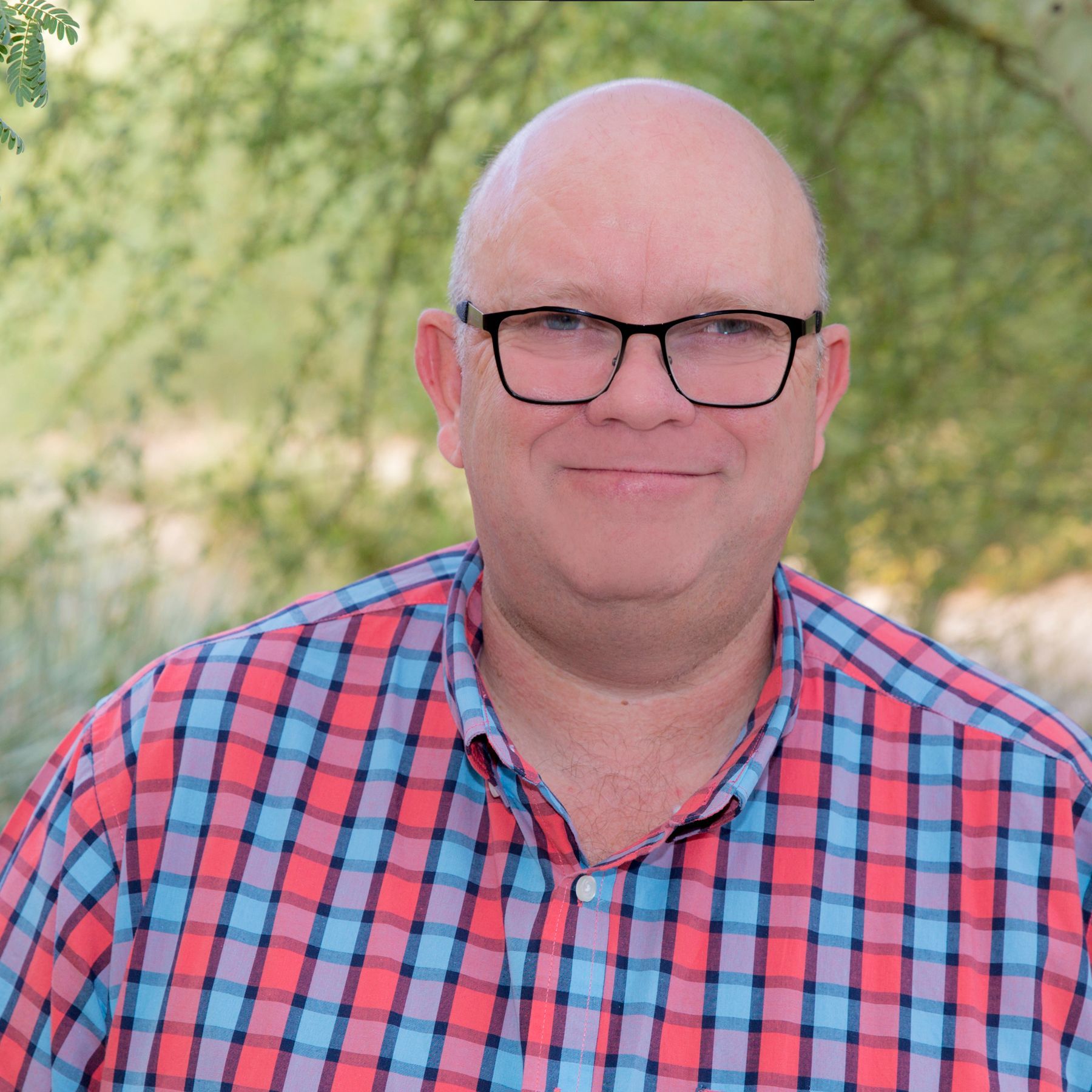 Bald man with glasses wearing a plaid shirt smiles outdoors in front of leafy background.