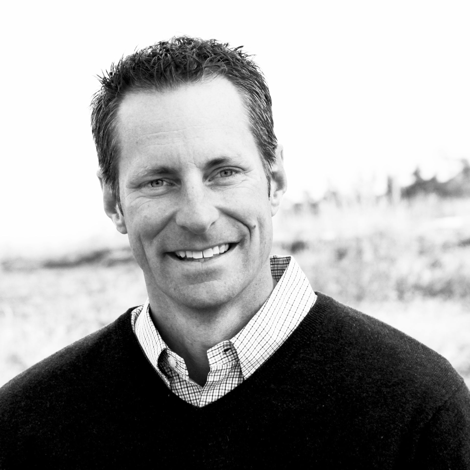 Man smiling, wearing a sweater over a collared shirt, outdoors with blurred background.