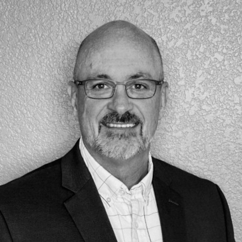 Man wearing glasses, blazer, and open-collar shirt smiles at the camera. Black and white photo.