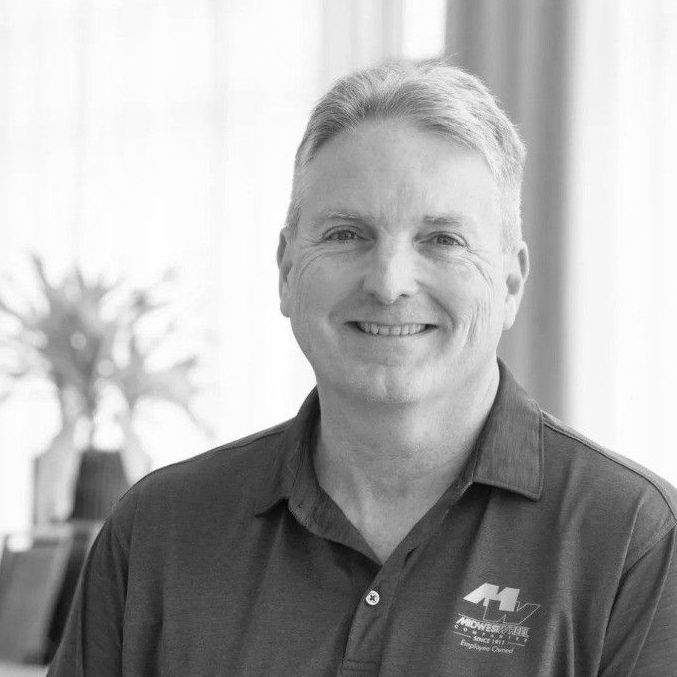 Man with graying hair smiles, wearing a polo shirt with a logo, in an office setting.