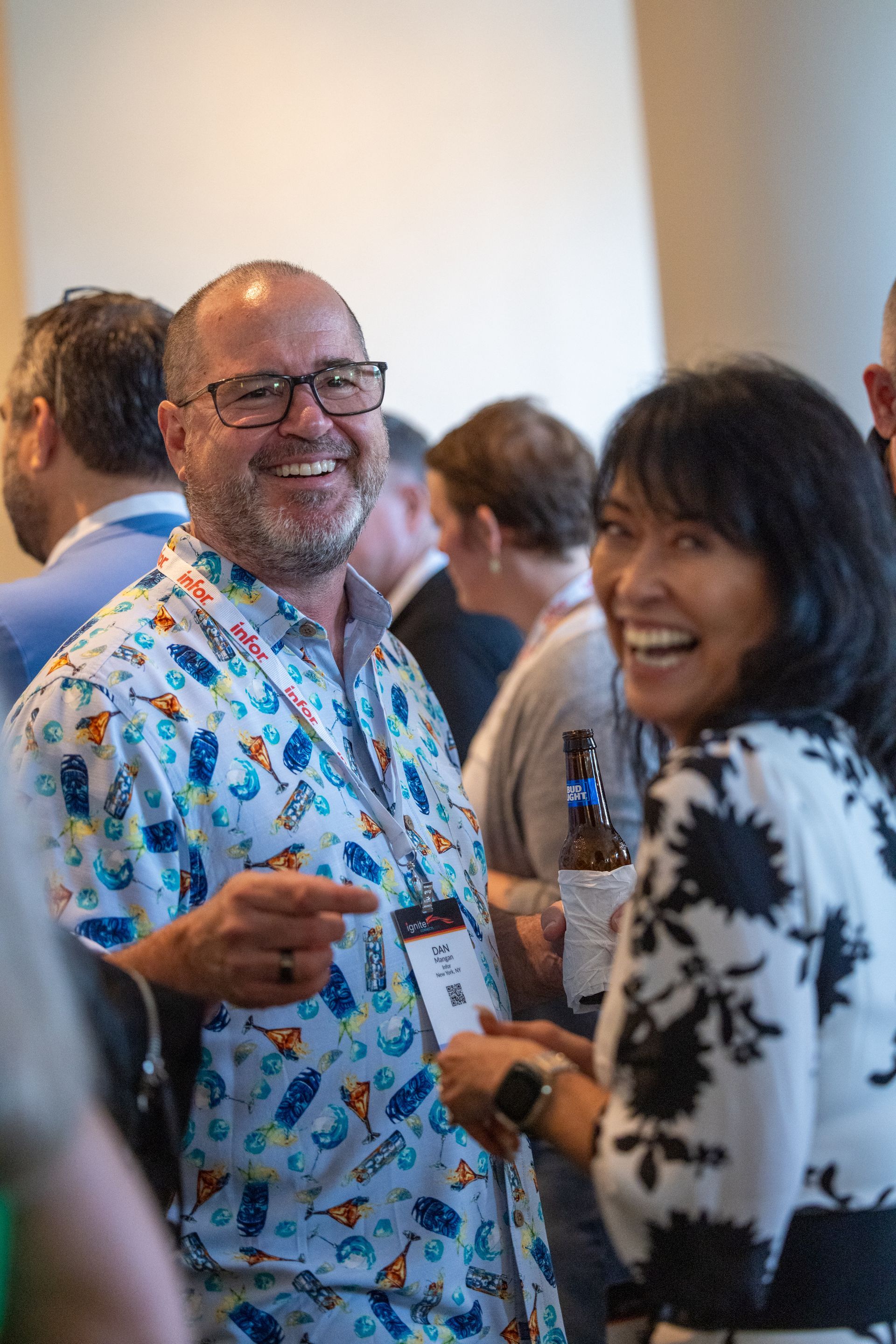 Man in patterned shirt points, smiling at woman in floral dress. People in background.